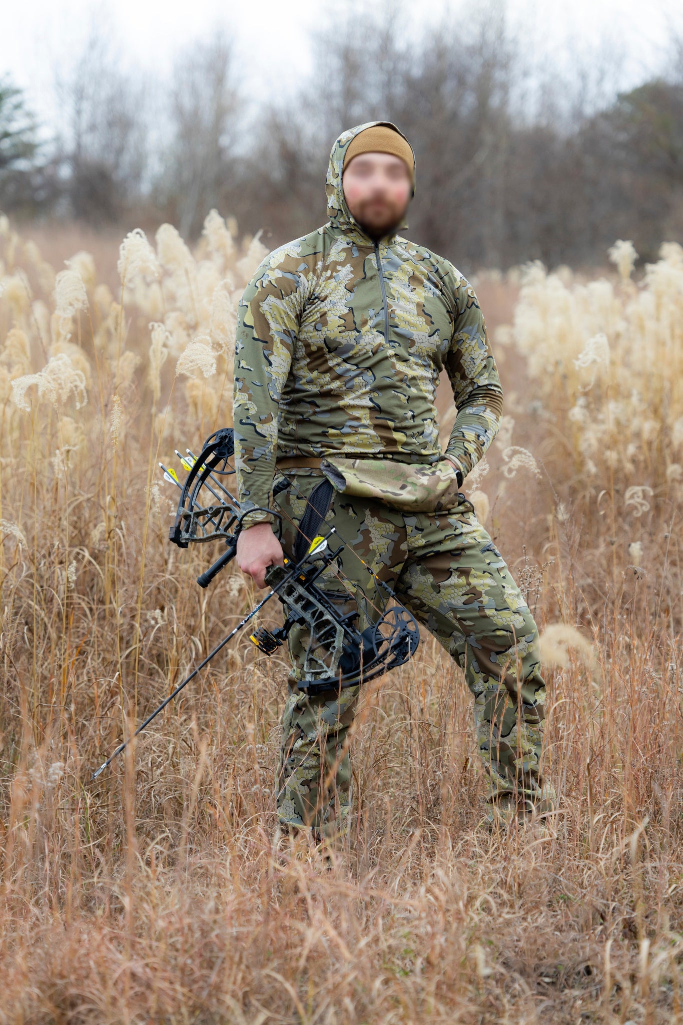 Hunter in Sitka camo and tan beanie carrying compound bow with Combat Effective hand warmer muff